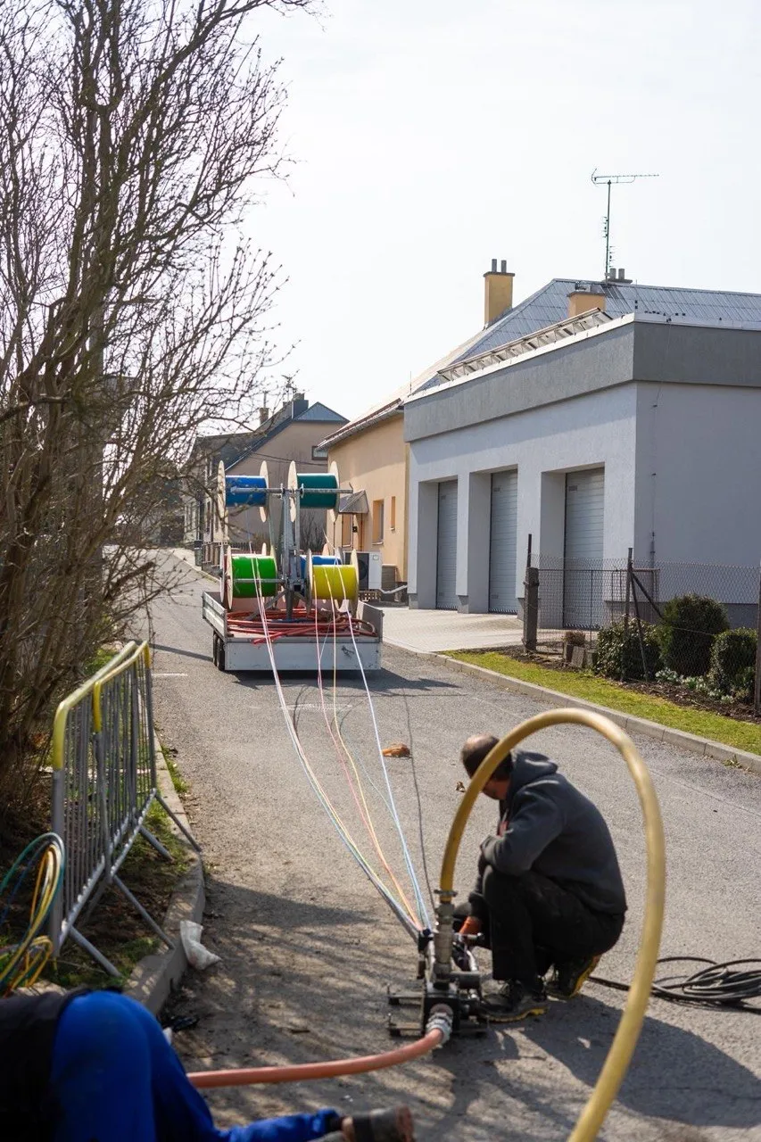 Technician at a fibre-blowing machine on a village street, with cable reels on a trailer in the background