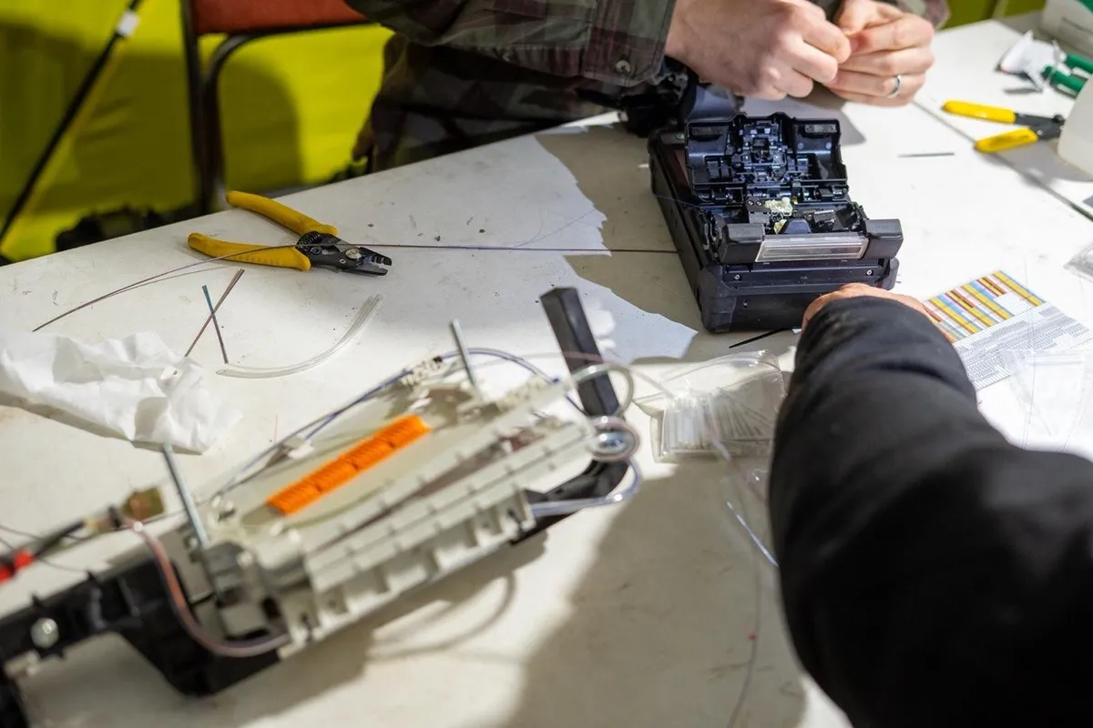 Hands at a fusion splicer, with an open splice tray and fibre tools on the table