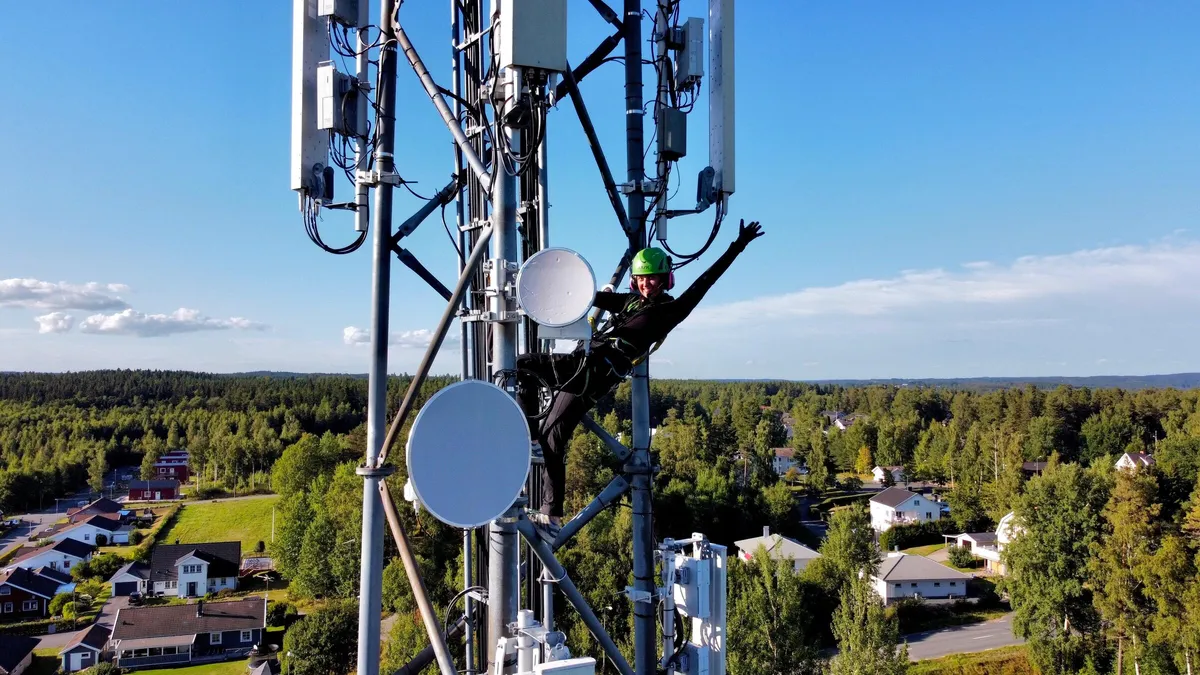 Rigger waving from a lattice tower carrying sector antennas and microwave dishes, with a village and forest below